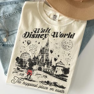 Peut inclure: T-shirt cr&egrave;me avec un motif noir du ch&acirc;teau de Walt Disney World, Mickey Mouse et des ballons. Le texte "Walt Disney World" et "The happiest place on earth" est imprim&eacute; sur le t-shirt.