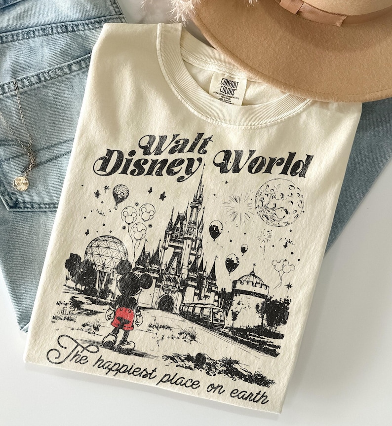 Peut inclure: T-shirt cr&egrave;me avec un motif noir du ch&acirc;teau de Walt Disney World, Mickey Mouse et des ballons. Le texte "Walt Disney World" et "The happiest place on earth" est imprim&eacute; sur le t-shirt.