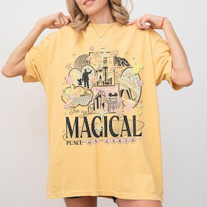 May include: Mustard yellow t-shirt with a colourful graphic design featuring a castle, Mickey Mouse, and the text "The Most Magical Place on Earth." The shirt has short sleeves and a relaxed fit.
