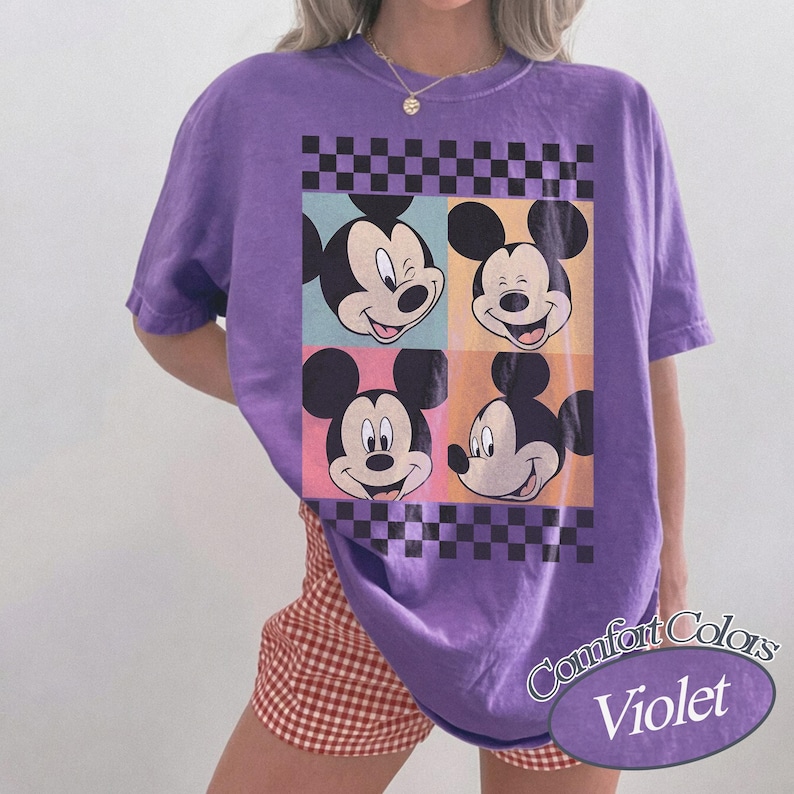May include: A violet Comfort Colors t-shirt featuring a graphic of Mickey Mouse's face in four different expressions. The design includes a checkered border and colorful squares. The shirt is a relaxed fit.