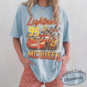 May include: Light blue t-shirt featuring a graphic of Lightning McQueen from the Cars movie. The design includes the text "Lightning 95 McQueen" in orange and yellow. The shirt is a Comfort Colors Chambray.
