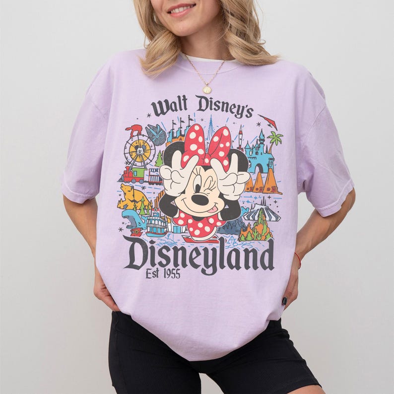 May include: A light purple t-shirt featuring a graphic of Minnie Mouse with the text "Walt Disney's Disneyland Est 1955". The graphic includes images of a Ferris wheel, a boat, and a castle.