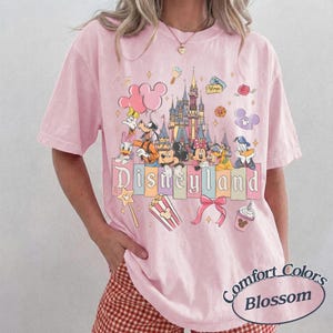 May include: A light pink t-shirt with a Disneyland graphic featuring Mickey Mouse, Minnie Mouse, Donald Duck, and Goofy. The shirt has a Comfort Colors Blossom label.