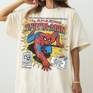 以下が含まれることがあります： 赤と青のスパイダーマンのコミックブックグラフィックが特徴のクリーム色のTシャツ。「The Amazing Spider-Man」の文字が大きく表示されています。吹き出しや「ZAP！」、「POW！」などのコミック要素も含まれています。