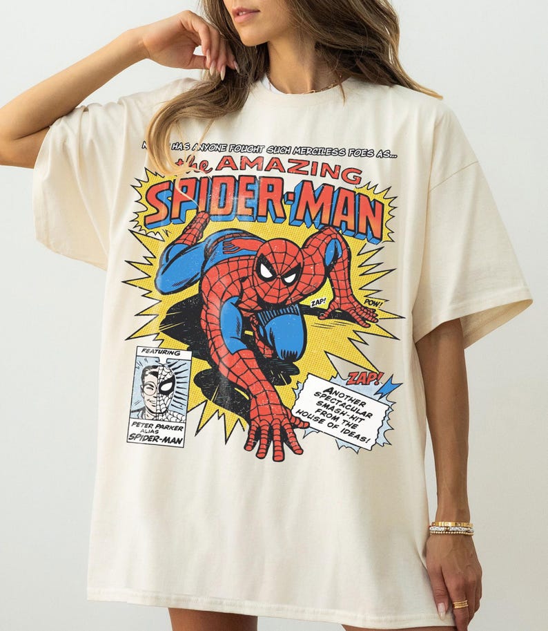 以下が含まれることがあります： 赤と青のスパイダーマンのコミックブックグラフィックが特徴のクリーム色のTシャツ。「The Amazing Spider-Man」の文字が大きく表示されています。吹き出しや「ZAP！」、「POW！」などのコミック要素も含まれています。