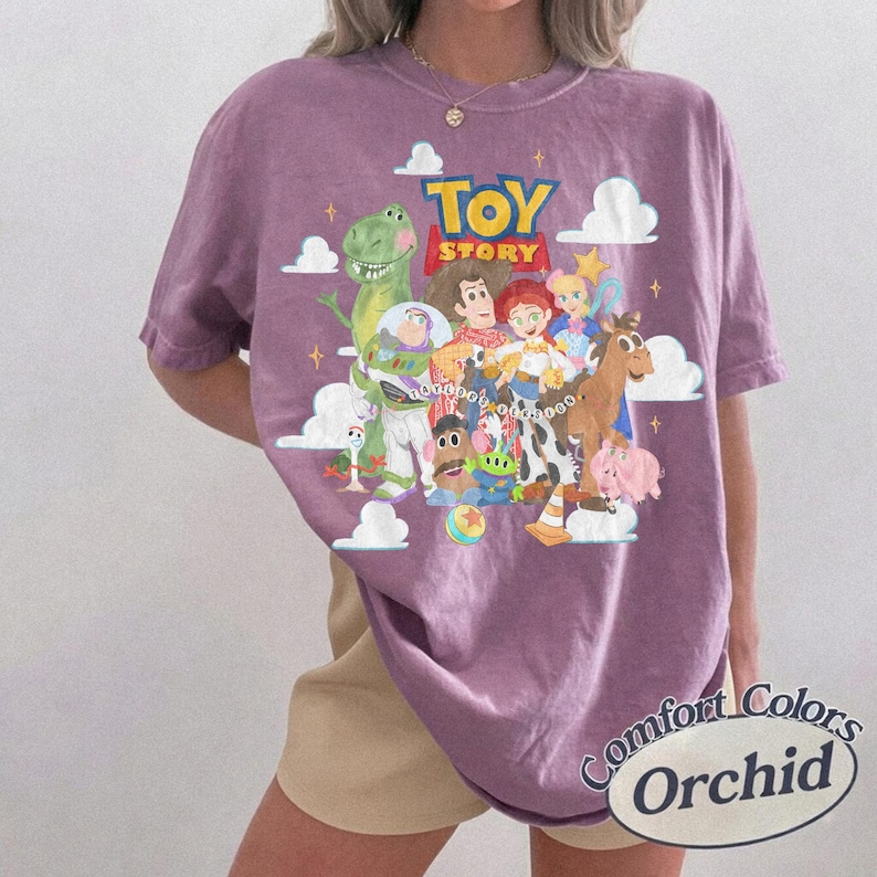 May include: A mauve crew neck t-shirt featuring a colorful graphic print of Toy Story characters including Woody, Buzz Lightyear, Rex, Jessie, and others, with the text "TOY STORY" in bold yellow and red letters surrounded by white clouds and stars.