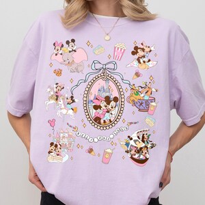 May include: A light purple t-shirt featuring a collage of Disney characters and icons. The design includes Mickey and Minnie Mouse, Dumbo, and the text "BEST DAY EVER". The shirt has a crew neck and short sleeves.