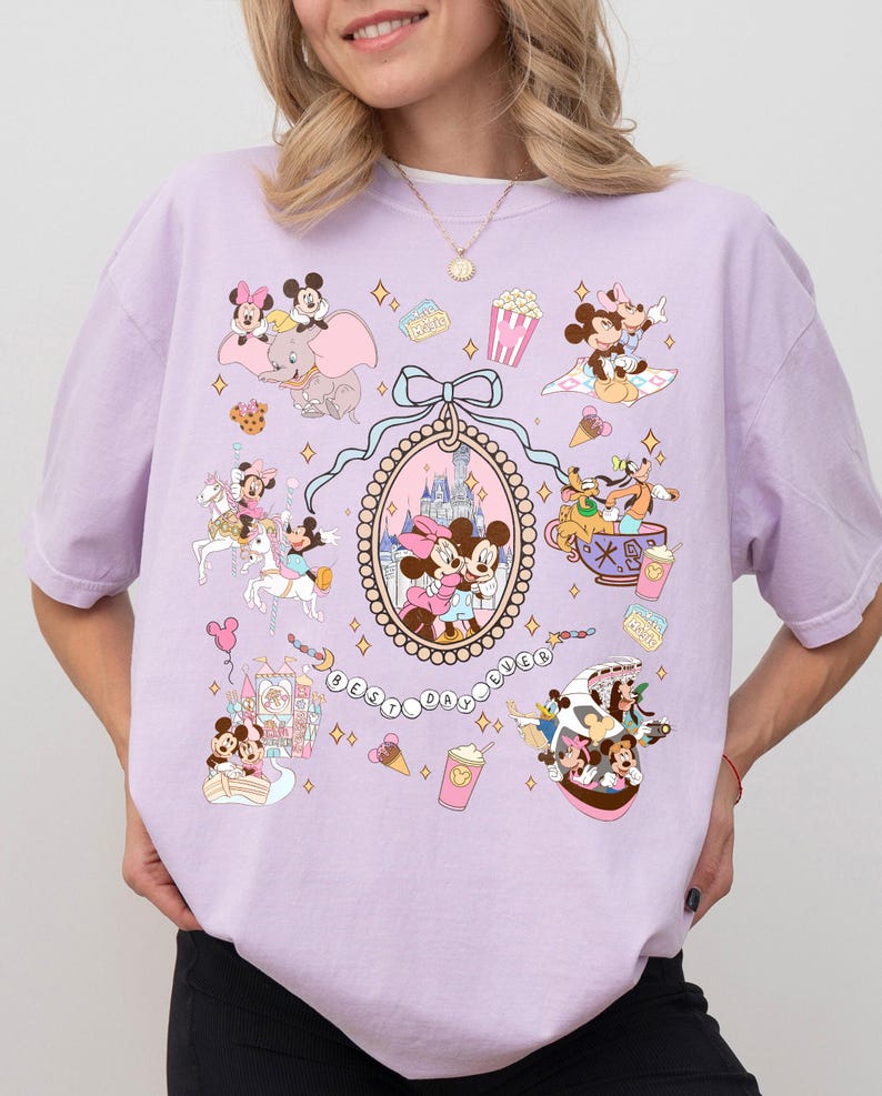 May include: A light purple t-shirt featuring a collage of Disney characters and icons. The design includes Mickey and Minnie Mouse, Dumbo, and the text "BEST DAY EVER". The shirt has a crew neck and short sleeves.