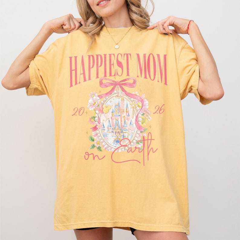 May include: Mustard yellow t-shirt with the text "Happiest Mom" in pink script. A pink bow and floral design surrounds a castle illustration. The numbers "20" and "26" flank the castle, with "on Earth" below.