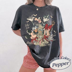 May include: Dark grey Comfort Colors t-shirt with a graphic of Disney characters in mummy costumes. The design includes Mickey Mouse, Minnie Mouse, Donald Duck, Daisy Duck, Goofy, and Pluto, with their names and signatures. The shirt has a relaxed fit.