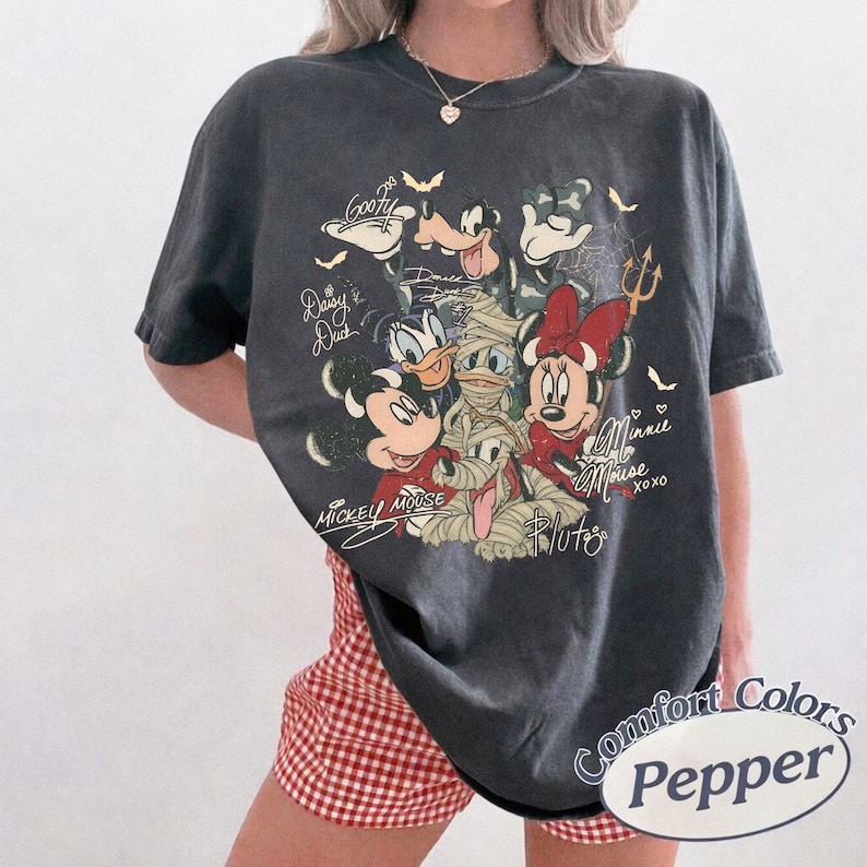 May include: Dark grey Comfort Colors t-shirt with a graphic of Disney characters in mummy costumes. The design includes Mickey Mouse, Minnie Mouse, Donald Duck, Daisy Duck, Goofy, and Pluto, with their names and signatures. The shirt has a relaxed fit.