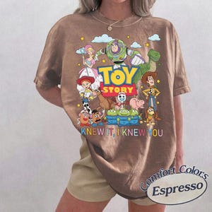 May include: A brown short-sleeve t-shirt featuring a colorful graphic of Toy Story characters including Buzz Lightyear, Woody, Jessie, and the Aliens. The text reads TOY STORY and I KNEW IT, I KNEW YOU. The shirt has a crew neckline and a relaxed fit.