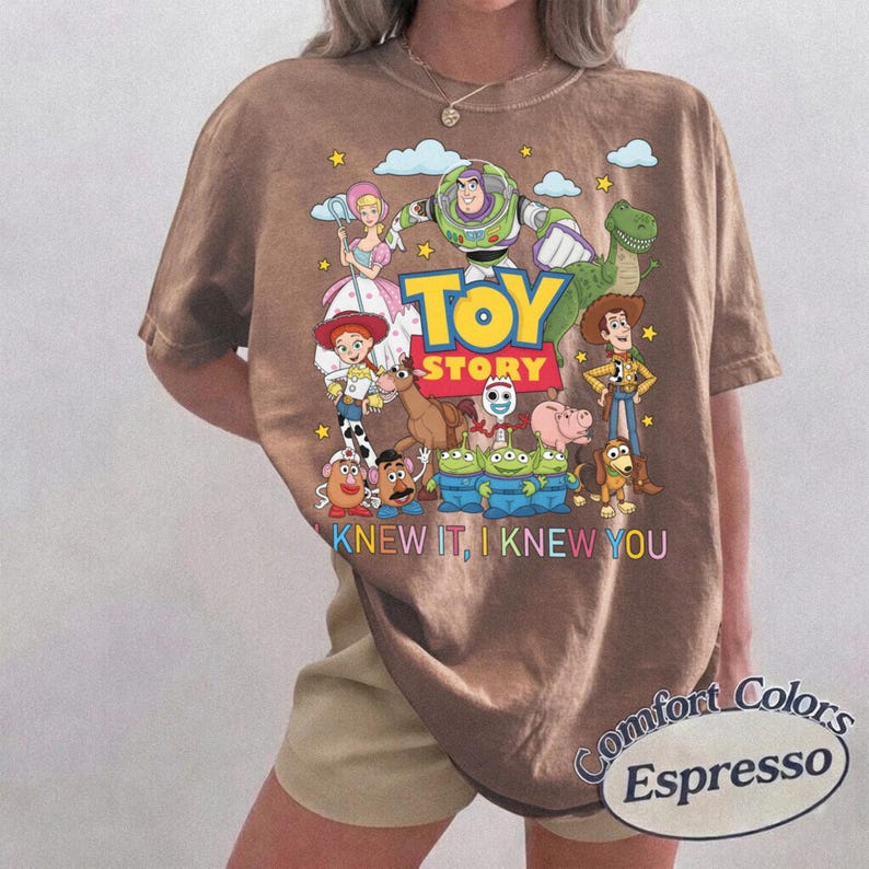 May include: A brown short-sleeve t-shirt featuring a colorful graphic of Toy Story characters including Buzz Lightyear, Woody, Jessie, and the Aliens. The text reads TOY STORY and I KNEW IT, I KNEW YOU. The shirt has a crew neckline and a relaxed fit.