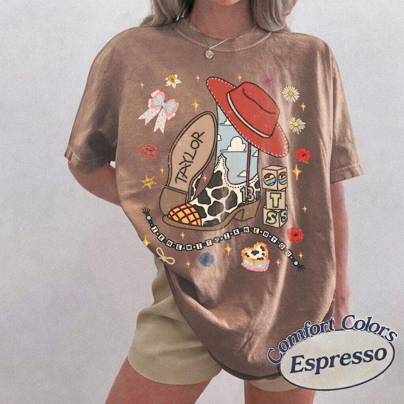 K&ouml;nnte beinhalten: Ein braunes Kurzarm-T-Shirt mit einem Grafikdruck aus einem Cowboystiefel mit Kuhmuster, einem roten Hut, einer Schleife, Blumen und Bl&ouml;cken mit dem Text Taylor, 13 und T S. Eine Perlenkette mit Text umgibt das zentrale Design auf dem Stoff.
