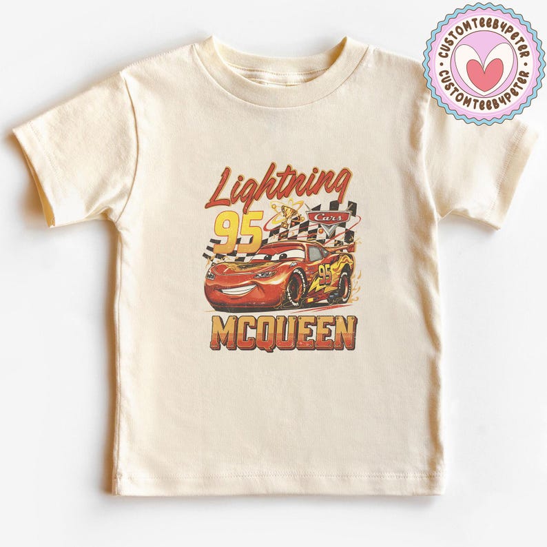 May include: Cream-colored t-shirt featuring a graphic of Lightning McQueen from the Cars movie. The design includes the text "Lightning 95 McQueen" in red and yellow, with a checkered flag and flames.