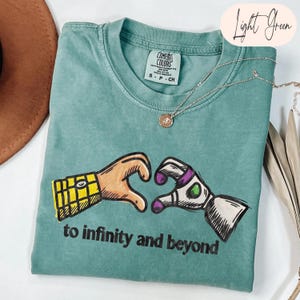 May include: A light green short-sleeve t-shirt featuring a graphic of two hands forming a heart shape, one resembling a yellow plaid shirt sleeve and the other a white and purple space suit glove, with the text to infinity and beyond below.