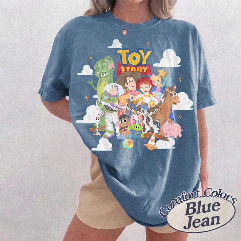 May include: A blue short-sleeve t-shirt featuring a graphic print of Toy Story characters including Woody, Buzz Lightyear, Rex, and Jessie. The design includes white clouds, yellow stars, and the red and yellow Toy Story logo text.
