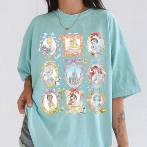 Pu&ograve; includere: Una maglietta azzurra con ritratti incorniciati di principesse animate. Ogni ritratto &egrave; circondato da dettagli floreali e fiocchi. Il design include un castello, una scarpetta di cristallo e una corona.