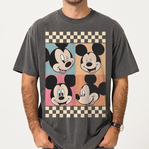 May include: Dark gray t-shirt featuring a colorful graphic of Mickey Mouse's face in four squares. The squares are in blue, orange, pink, and beige, with a checkered border. The shirt has a vintage, worn look.