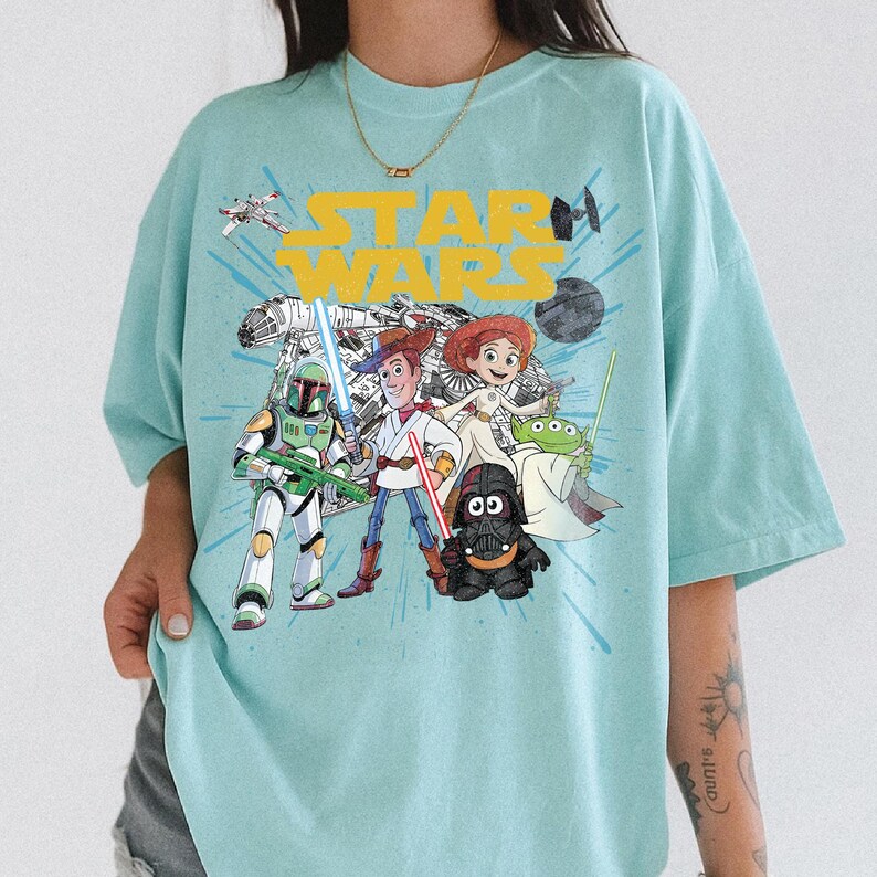 Puede incluir: Camiseta azul claro con un gr&aacute;fico de personajes de Star Wars, incluyendo Darth Vader, Woody y Buzz Lightyear. Las palabras "STAR WARS" son amarillas. La camiseta tiene un corte holgado y es de material suave.