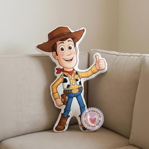 Peut inclure: Un coussin en forme du personnage Woody du film d'animation Toy Story. Woody porte un chapeau de cowboy marron, une chemise &agrave; carreaux jaune, un jean bleu et des bottes marron. Il fait un signe de pouce.