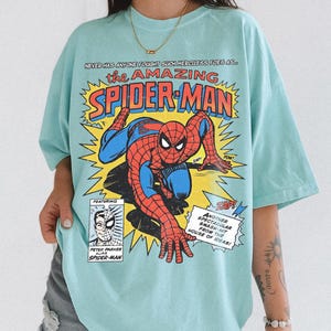 以下が含まれることがあります： 水色のTシャツ。赤と青のスパイダーマンのコミックブックグラフィックが特徴です。「the AMAZING SPIDER-MAN」の文字は黄色と赤で、コミックブックのテキストとイラストが描かれています。