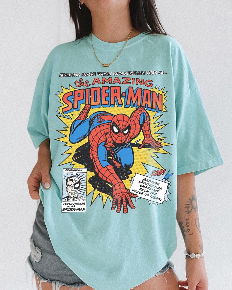 以下が含まれることがあります： 水色のTシャツ。赤と青のスパイダーマンのコミックブックグラフィックが特徴です。「the AMAZING SPIDER-MAN」の文字は黄色と赤で、コミックブックのテキストとイラストが描かれています。