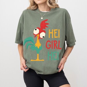 Peut inclure: T-shirt vert olive avec un dessin de coq de dessin anim&eacute; aux accents rouges, jaunes et turquoise. Le texte "HEI GIRL HEI" est imprim&eacute; en jaune, rouge et turquoise. Le t-shirt a des manches courtes.