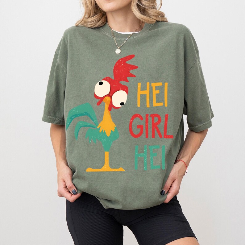 Peut inclure: T-shirt vert olive avec un dessin de coq de dessin anim&eacute; aux accents rouges, jaunes et turquoise. Le texte "HEI GIRL HEI" est imprim&eacute; en jaune, rouge et turquoise. Le t-shirt a des manches courtes.