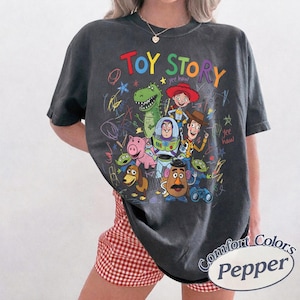 May include: Dark gray t-shirt featuring a colorful "Toy Story" graphic with cartoon characters like Buzz Lightyear, Woody, and Rex. The shirt has a crew neck and short sleeves. The text "Comfort Colors Pepper" is printed on the bottom right.