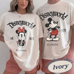 May include: Ivory-colored t-shirts with the text "Disneyworld" and the year "1971". One shirt features Minnie Mouse, the other Mickey Mouse. Both shirts have "Florida" printed below the characters. The shirts are paired with red and white checkered shorts.