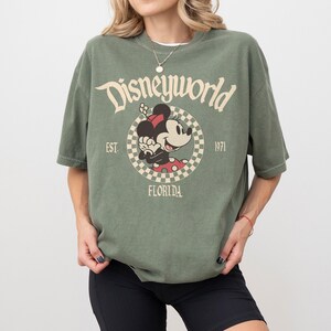 May include: Olive green t-shirt with a Disneyworld graphic featuring Minnie Mouse. The design includes the text "Disneyworld", "Est. 1971", and "Florida". The shirt has short sleeves and a relaxed fit.