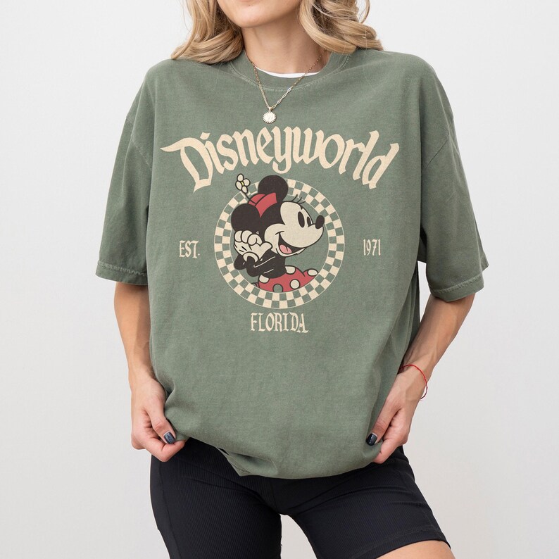 May include: Olive green t-shirt with a Disneyworld graphic featuring Minnie Mouse. The design includes the text "Disneyworld", "Est. 1971", and "Florida". The shirt has short sleeves and a relaxed fit.