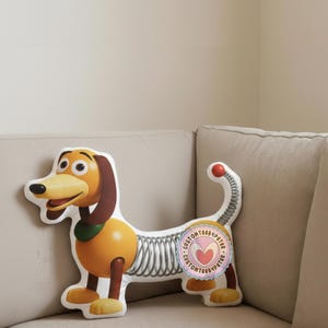 Peut inclure: Peluche en forme de chien avec un corps jaune, des oreilles marron et une queue rouge. Le jouet a un collier vert et un visage souriant. Un autocollant avec du texte est visible sur le c&ocirc;t&eacute;.
