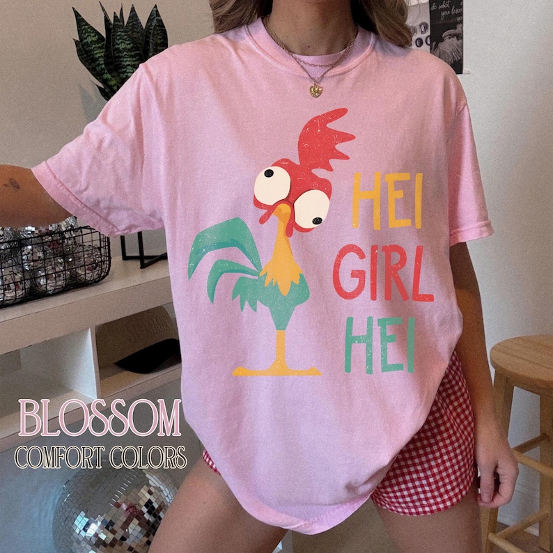 Peut inclure: T-shirt rose clair avec un dessin de coq de dessin anim&eacute; et le texte "HEI GIRL HEI" en jaune, rouge et vert. Le t-shirt porte &eacute;galement le texte "BLOSSOM COMFORT COLORS". Le t-shirt est port&eacute; avec un short &agrave; carreaux rouges et blancs.