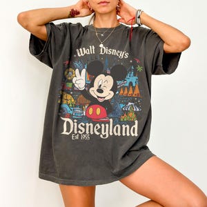 May include: Dark gray t-shirt featuring Mickey Mouse giving a peace sign in front of a colorful Disneyland graphic. The shirt has the text "Walt Disney's Disneyland Est 1955". The shirt is oversized and has short sleeves.