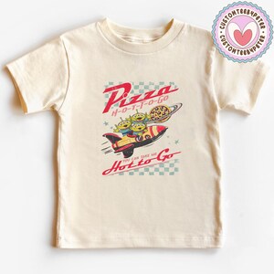 Pu&ograve; includere: T-shirt color crema con grafica in stile retr&ograve;. Il design include il testo "Pizza H-O-T-T-O-GO" con un razzo che trasporta tre alieni verdi e una pizza. &Egrave; presente anche la frase "You can take me Hot to Go".