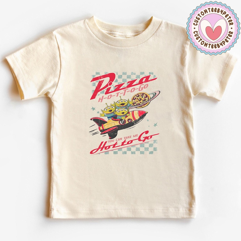 Pu&ograve; includere: T-shirt color crema con grafica in stile retr&ograve;. Il design include il testo "Pizza H-O-T-T-O-GO" con un razzo che trasporta tre alieni verdi e una pizza. &Egrave; presente anche la frase "You can take me Hot to Go".
