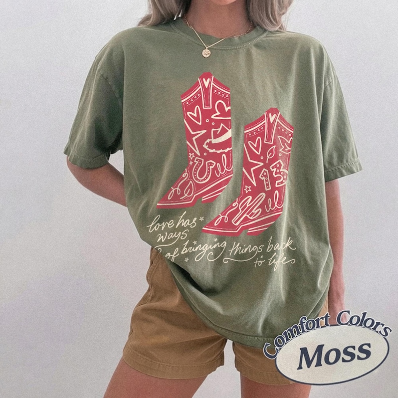 May include: An olive green short-sleeve t-shirt featuring a graphic of two red cowboy boots decorated with hearts, stars, and the number 13. Below the boots, the text reads, love has ways of bringing things back to life in white cursive script.