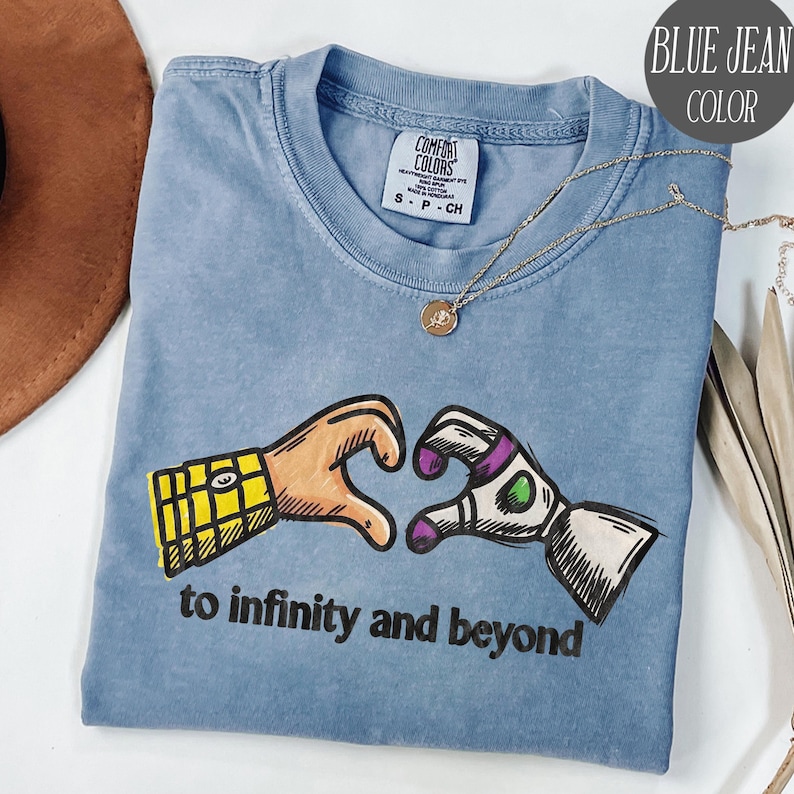 May include: A blue jean colored t-shirt featuring a graphic of two hands forming a heart shape, one resembling a yellow plaid shirt sleeve and the other a white and purple space suit sleeve, with the text to infinity and beyond printed below.