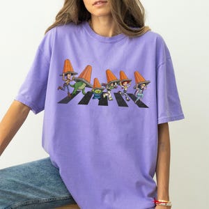 Puede incluir: Una camiseta de color lila con un gr&aacute;fico de personajes de dibujos animados cruzando una calle, cada uno con un cono de tr&aacute;fico naranja. El dise&ntilde;o es una parodia de la ic&oacute;nica portada del &aacute;lbum Abbey Road. La camiseta tiene mangas cortas y un corte holgado.