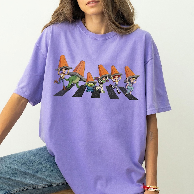 Puede incluir: Una camiseta de color lila con un gr&aacute;fico de personajes de dibujos animados cruzando una calle, cada uno con un cono de tr&aacute;fico naranja. El dise&ntilde;o es una parodia de la ic&oacute;nica portada del &aacute;lbum Abbey Road. La camiseta tiene mangas cortas y un corte holgado.