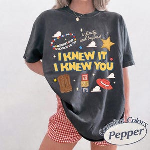 May include: A charcoal gray short-sleeve t-shirt featuring a graphic print with the text "I KNEW IT I KNEW YOU", "infinity and beyond", and "YOU'VE GOT A FRIEND IN ME". The design includes illustrations of a cowboy boot, a red hat, and alphabet blocks.