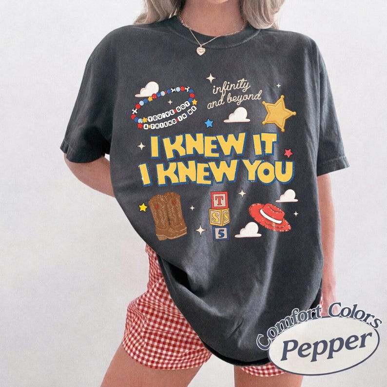 May include: A charcoal gray short-sleeve t-shirt featuring a graphic print with the text "I KNEW IT I KNEW YOU", "infinity and beyond", and "YOU'VE GOT A FRIEND IN ME". The design includes illustrations of a cowboy boot, a red hat, and alphabet blocks.