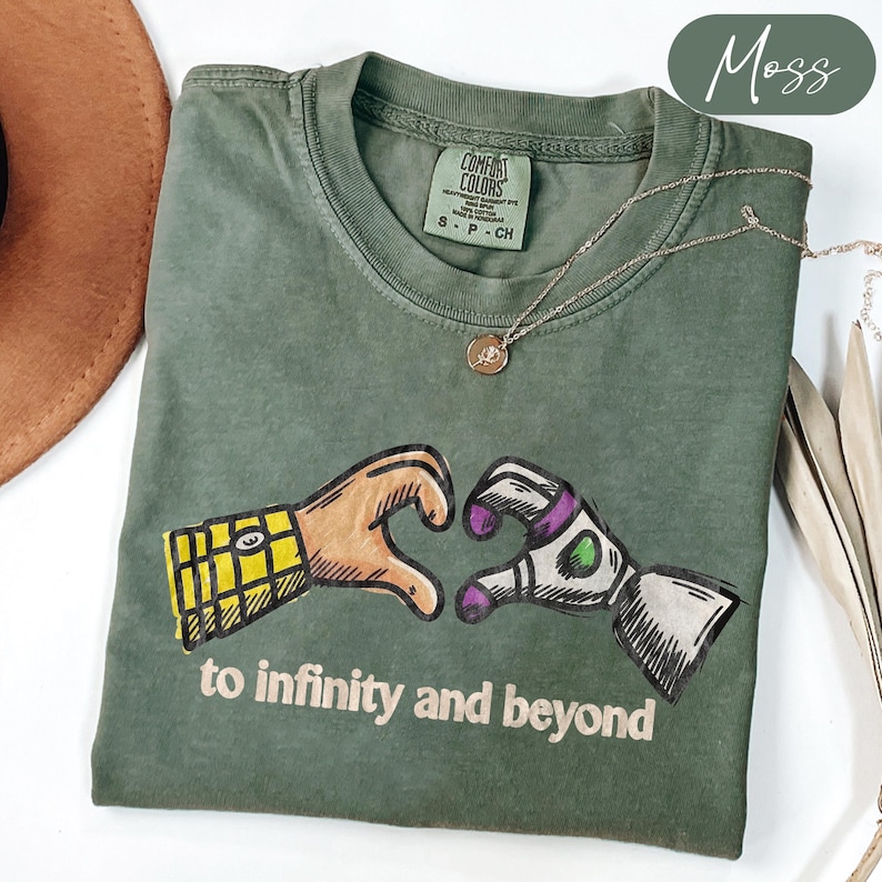 May include: A moss green t-shirt featuring a graphic of two hands forming a heart shape, one with a yellow plaid sleeve and the other with a white and purple robotic sleeve. The text "to infinity and beyond" is printed below the graphic.