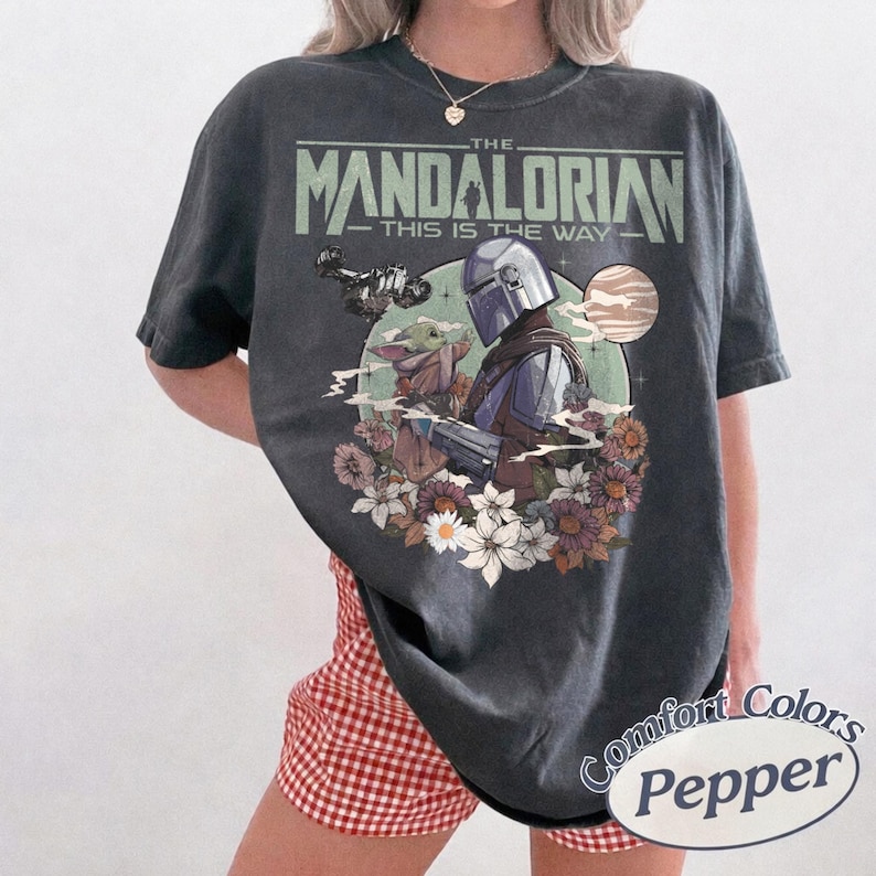 Może przedstawiać: Ciemnoszara koszulka z grafiką z "The Mandalorian". Projekt zawiera napis "The Mandalorian - This is the Way" nad ilustracją Mandalorianina i Grogu otoczonych kwiatami. Koszulka jest marki Comfort Colors.