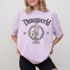 May include: A light purple t-shirt with the text "Disneyworld" in black script. The shirt features a circular graphic of Daisy Duck, with the text "EST. 1971 FLORIDA". The shirt is short-sleeved.