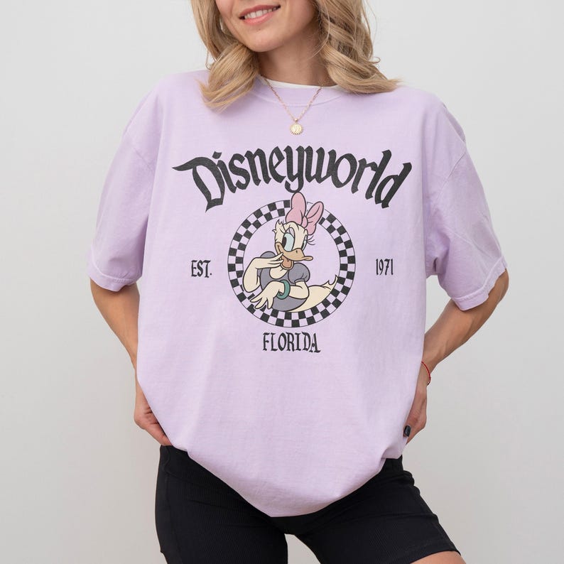 May include: A light purple t-shirt with the text "Disneyworld" in black script. The shirt features a circular graphic of Daisy Duck, with the text "EST. 1971 FLORIDA". The shirt is short-sleeved.