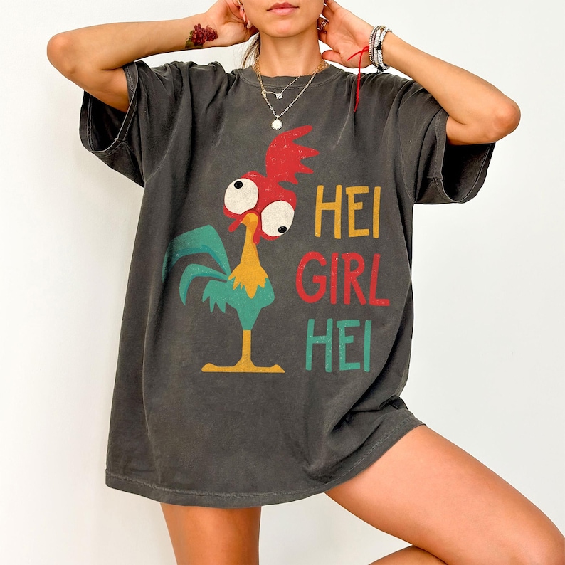 Peut inclure: T-shirt gris fonc&eacute; avec un coq de dessin anim&eacute; aux accents rouges, jaunes et turquoise. Le texte "HEI GIRL HEI" est imprim&eacute; en jaune, rouge et turquoise. Le t-shirt est ample et &agrave; manches courtes.