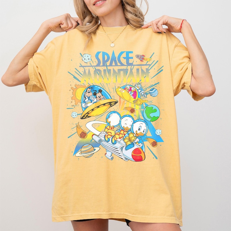 Pu&ograve; includere: T-shirt giallo senape con una grafica colorata di personaggi Disney a tema spaziale. Il design include Topolino, Paperino e il testo "SPACE MOUNTAIN". La maglietta ha una vestibilit&agrave; ampia.